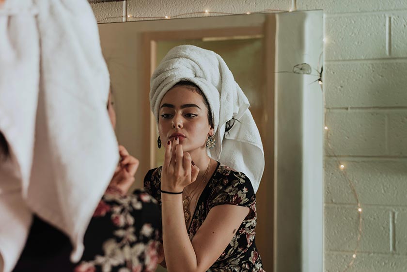 Junge Frau mit Handtuchturban schminkt sich im Badezimmerspiegel