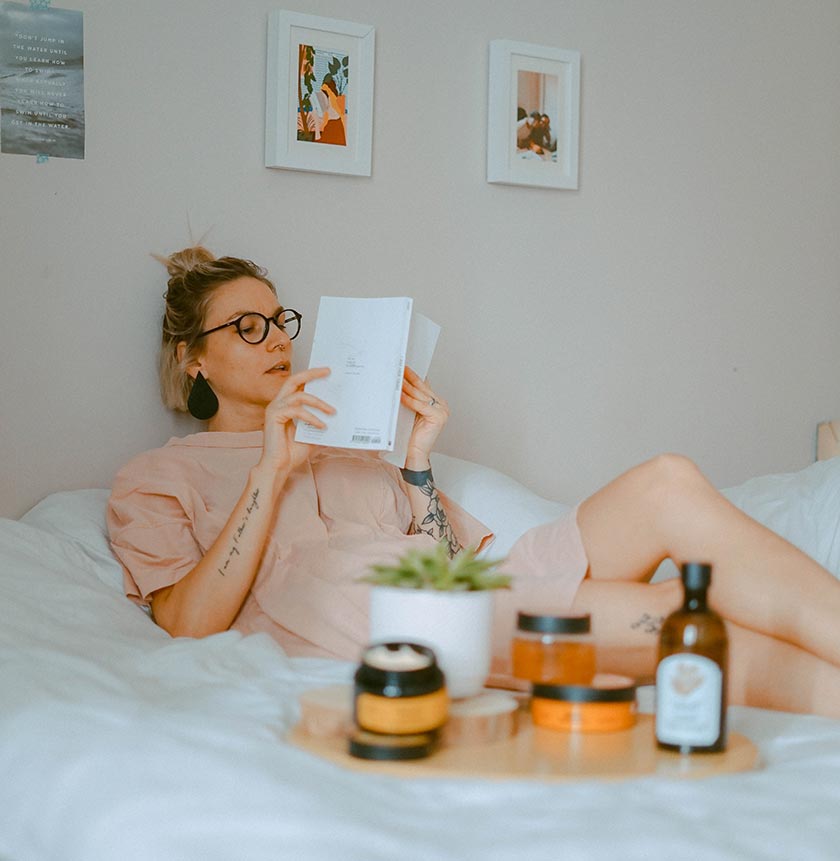 woman reading book on bed with skincare products and plant