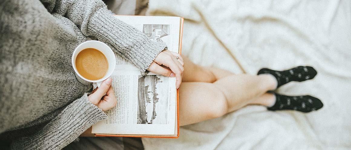 Frau sitzt mit Kaffee und Buch auf dem Bett und liest
