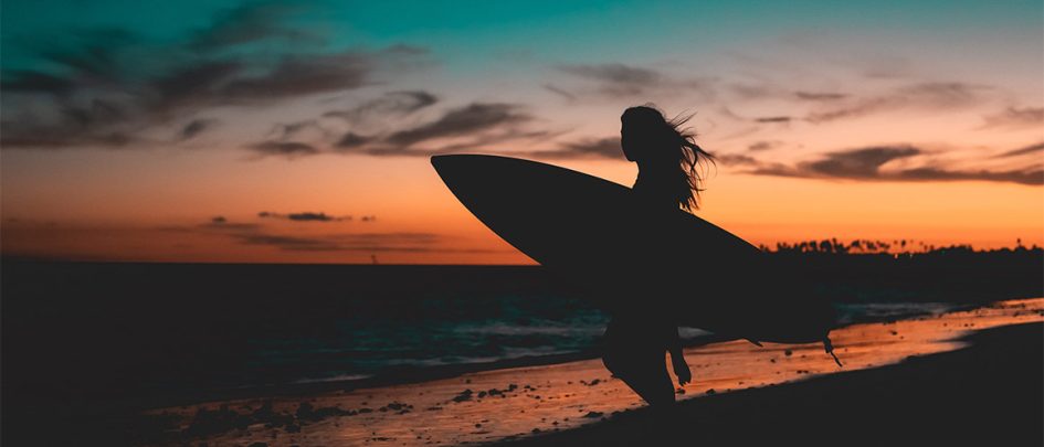 woman standing at the beach with a surfboard at sunset
